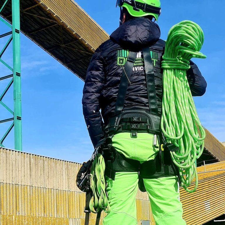 Cordiste de MP Ouest Services vu de dos, harnais complet et cordes sur l'épaule, au pied d'une structure industrielle